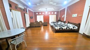 A hotel room with a wooden floor, a round dining table with chairs, a sofa, a bed with a patterned bedspread, and a ceiling light fixture. - Wisma Acidalia Syariah (Bandung)