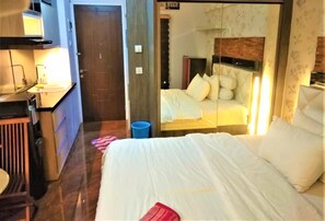 A hotel room with a bed, a desk with a lamp, a trash bin, and a door. - Apartemen Mutiara Bekasi By Veeroom (Bekasi)