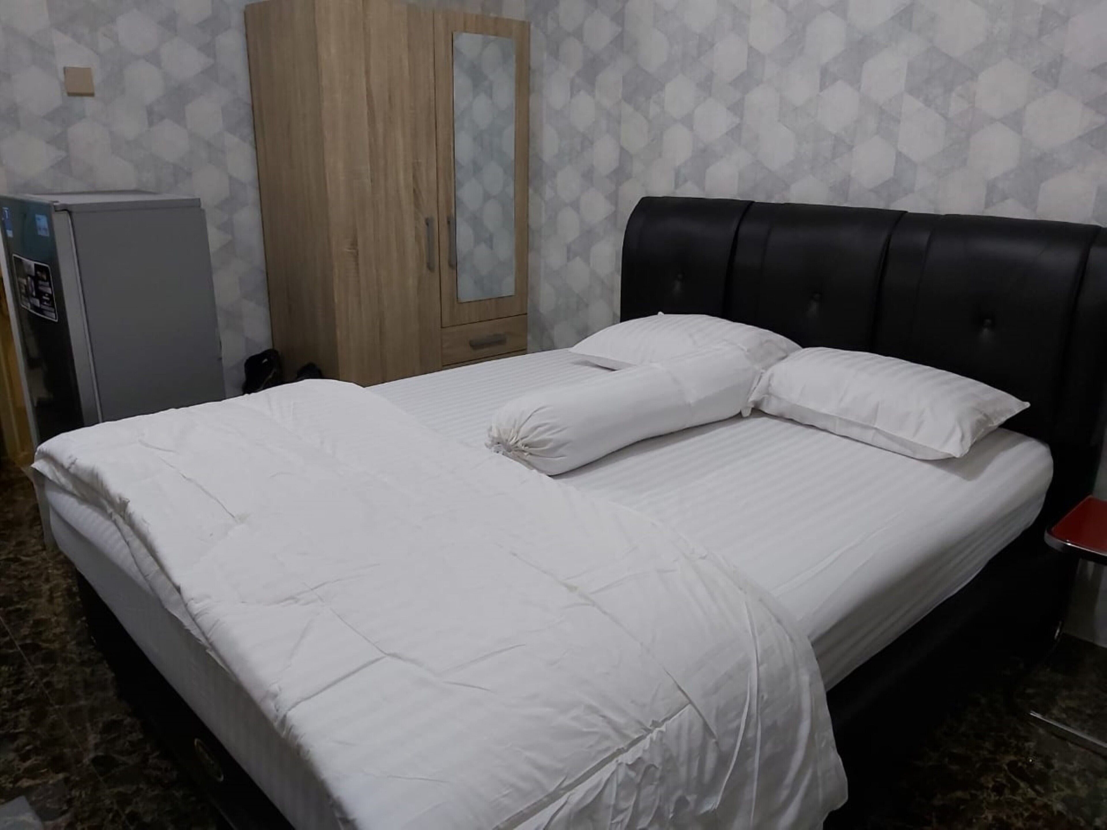 A neatly made bed with white linens and pillows, a wooden wardrobe with a mirror, and a small refrigerator.