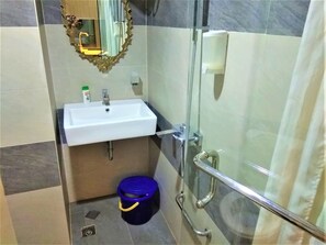 A bathroom with a white sink, a mirror with an ornate frame, a shower with a glass door, and a blue bucket on the floor. - Apartemen Mutiara Bekasi By Veeroom (Bekasi)