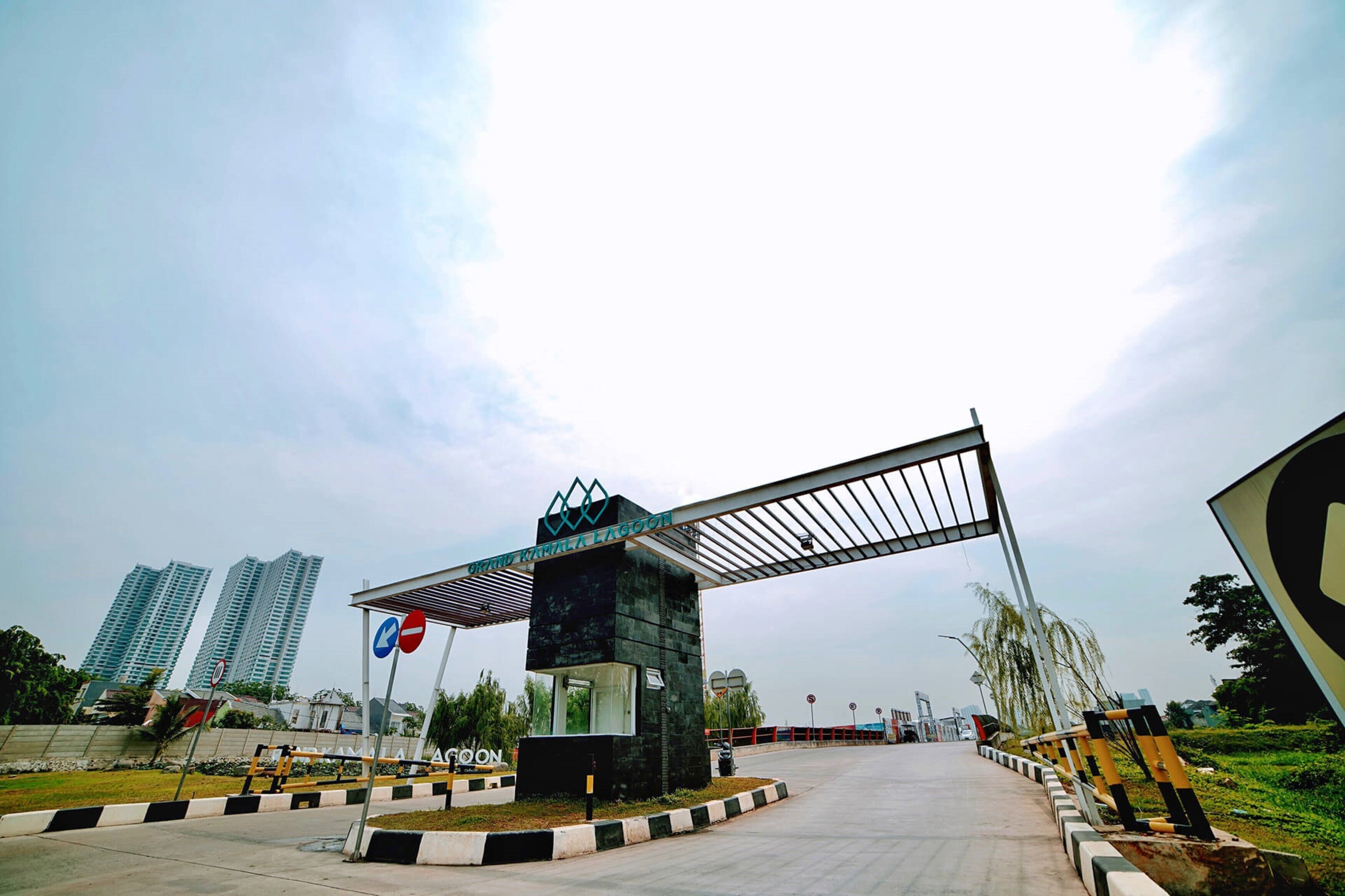 A gated entrance with a metal archway, a sign with a blue and red symbol, and tall buildings in the background.
