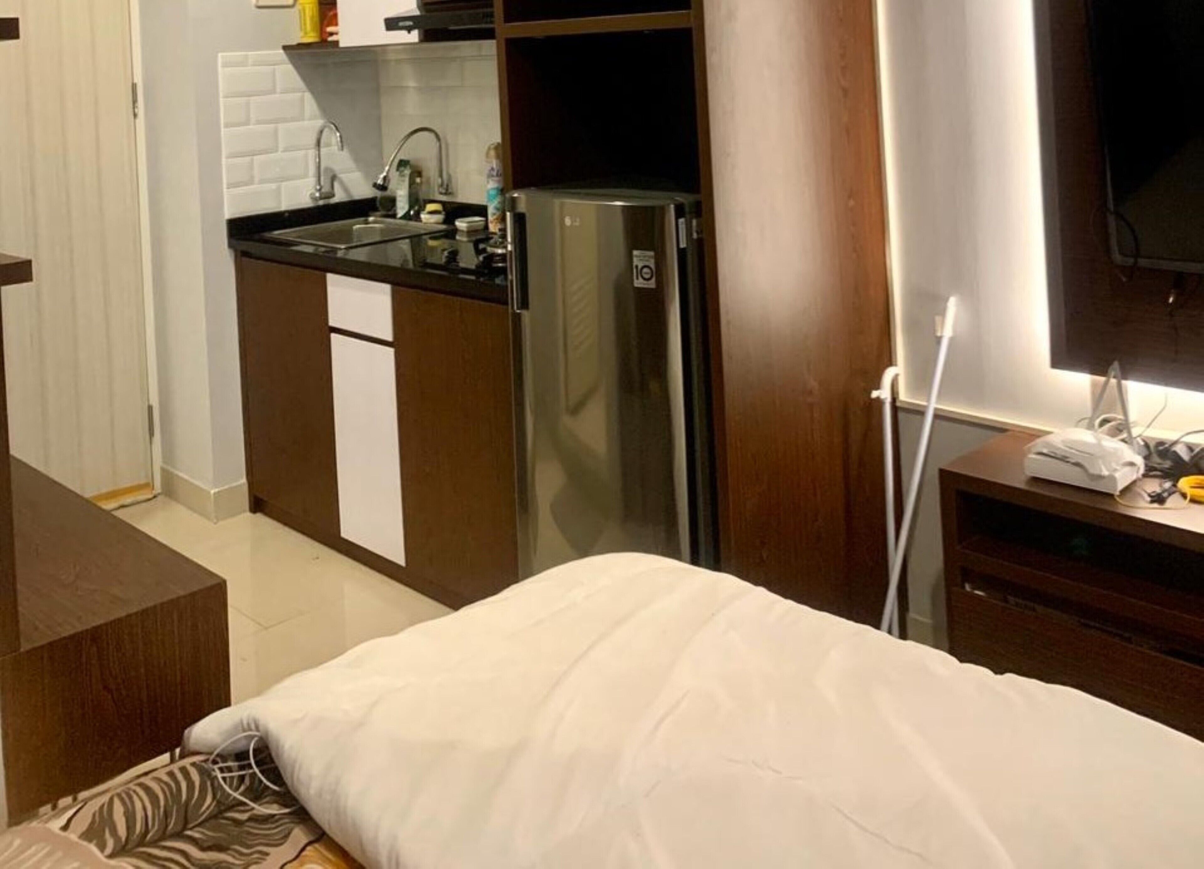 Quarto compacto com cama, cozinha compacta equipada com geladeira e televisão na parede.