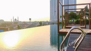 An infinity pool with a wooden deck and metal railings, overlooking a cityscape with high-rise buildings and a mosque.