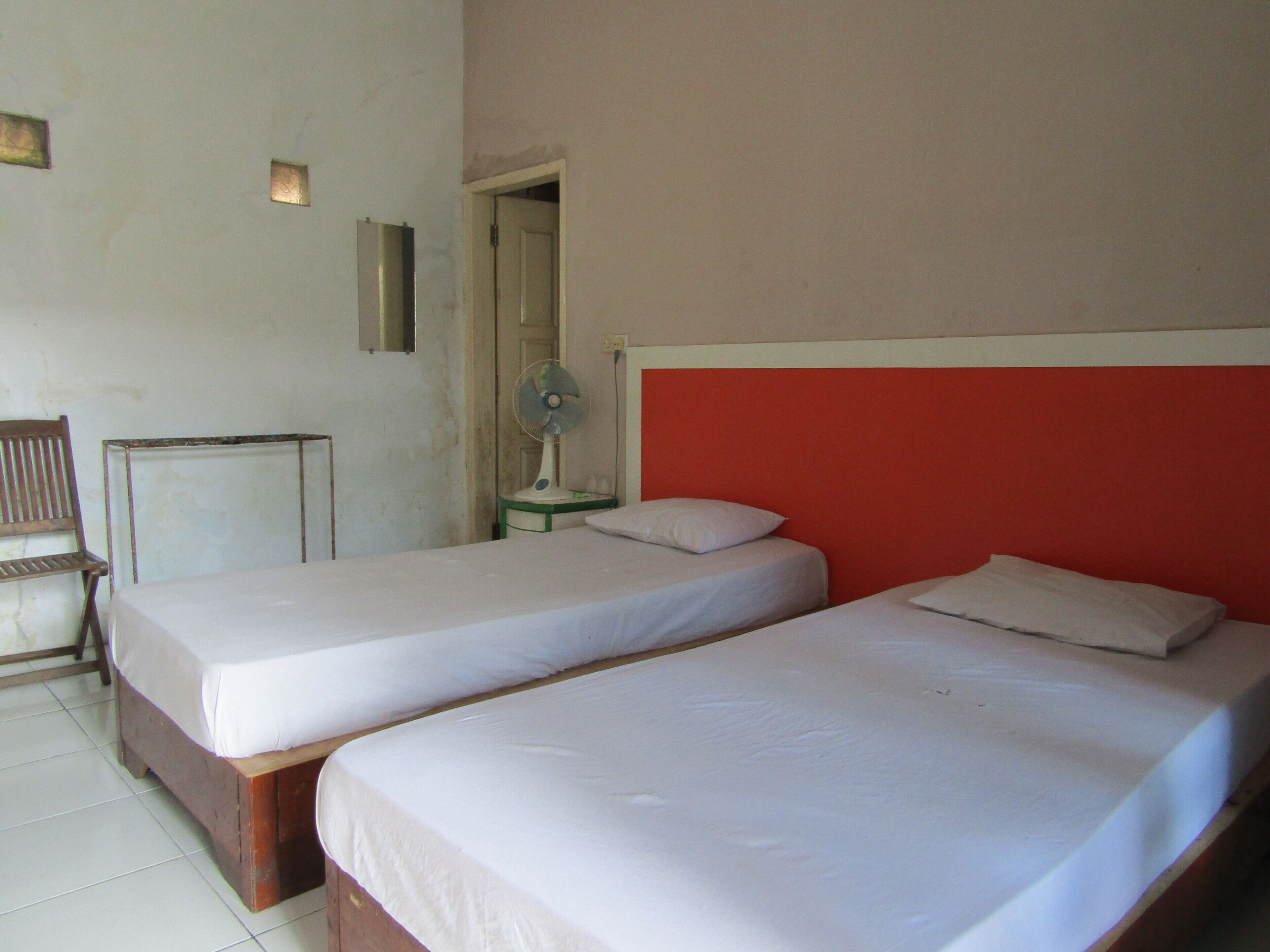 Ein Zimmer mit zwei Einzelbetten, einem Holzstuhl, einem kleinen Tisch und einem Wandventilator.