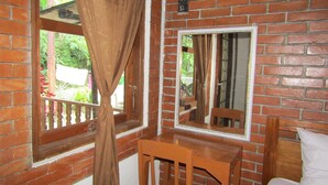 Quarto com parede de tijolos, cadeira de madeira, cama, janela com cortinas e um espelho.
