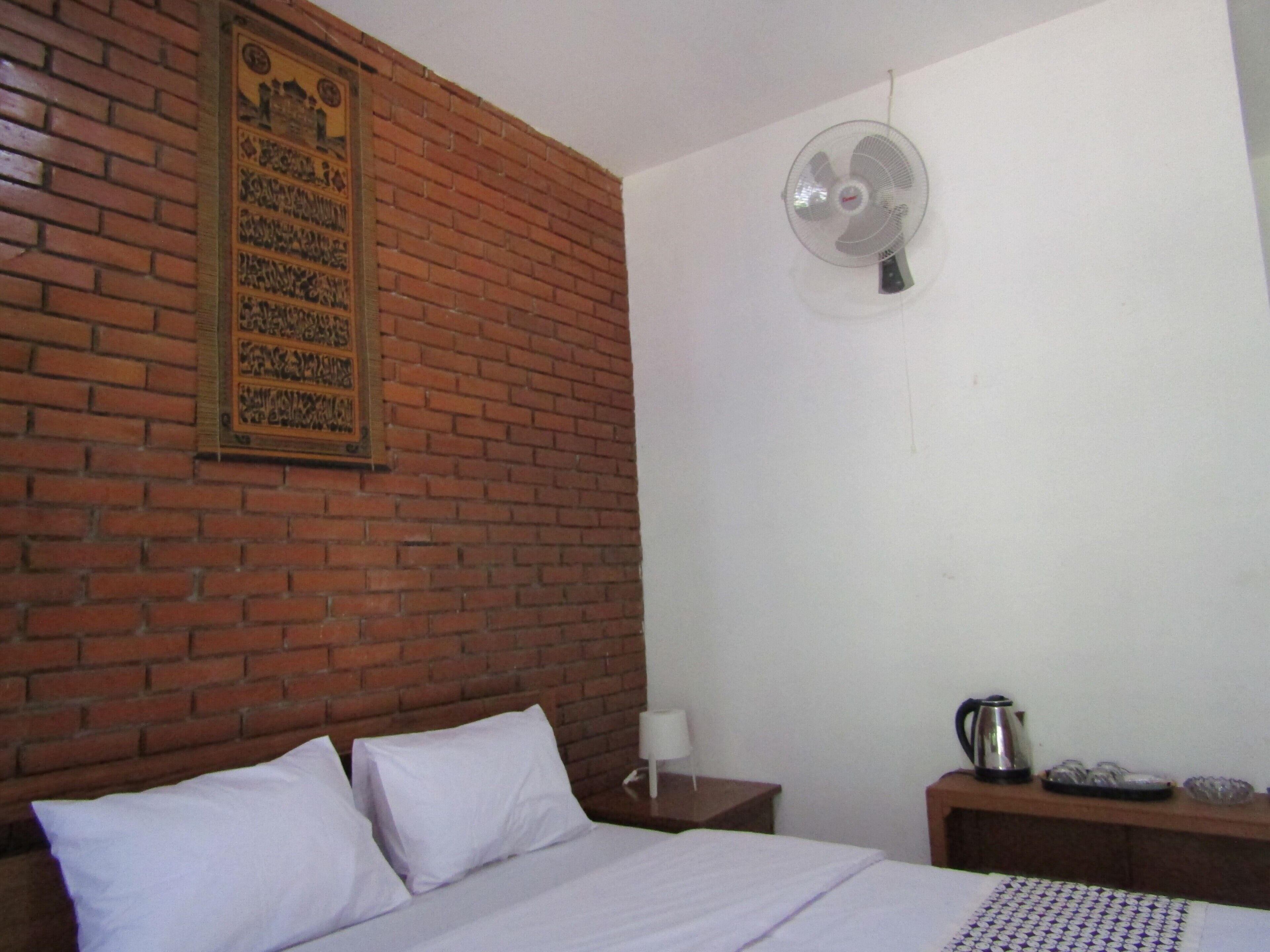 A room with a brick wall, a bed with white bedding, a wall-mounted fan, a nightstand with a lamp, and a small table with a kettle and cups.