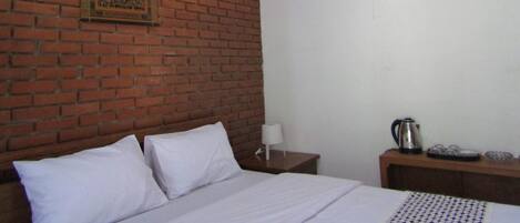 A room with a brick wall, a bed with white bedding, a wall-mounted fan, a nightstand with a lamp, and a small table with a kettle and cups.