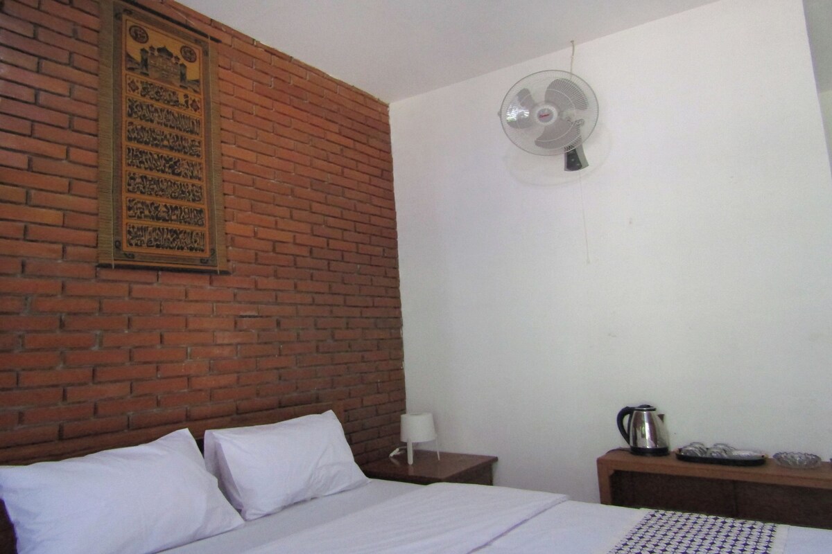 A hotel room with a brick wall, a bed with white linens, a bedside table with a lamp and a kettle, and a wall-mounted fan.
