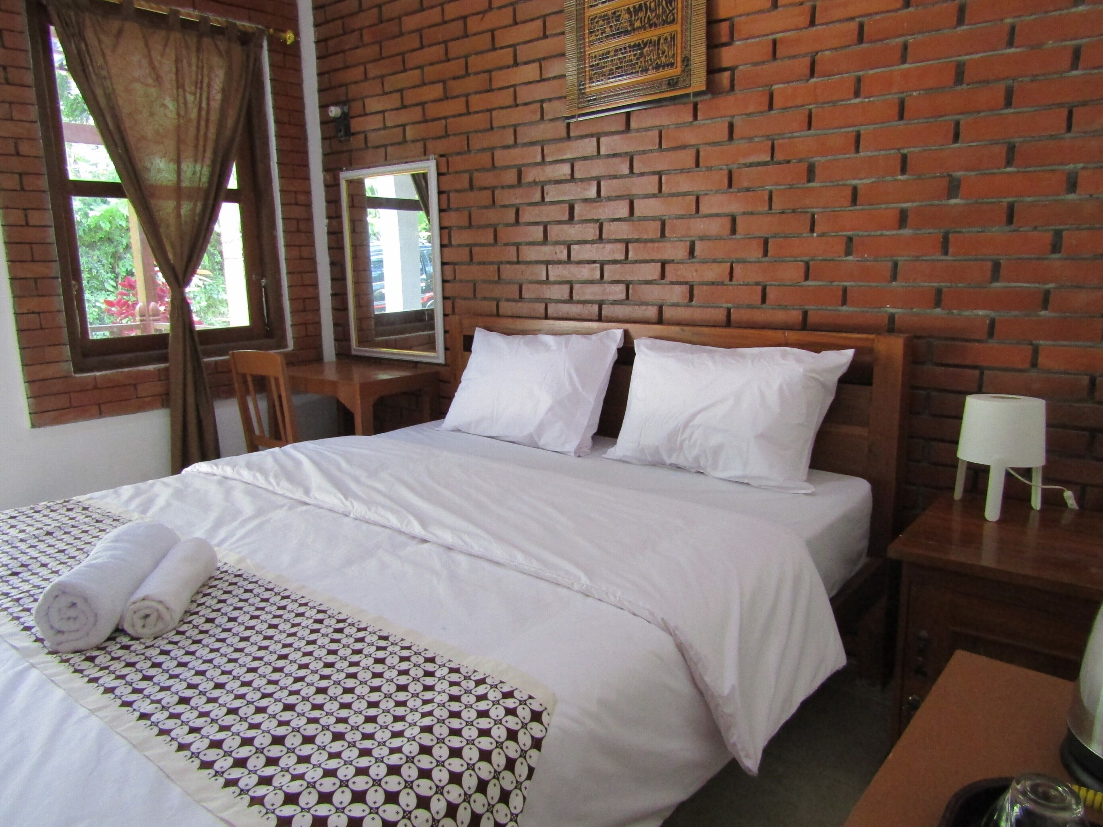 A hotel room with a brick wall, a bed with white bedding, a wooden headboard, a bedside table with a lamp, and a window with curtains.