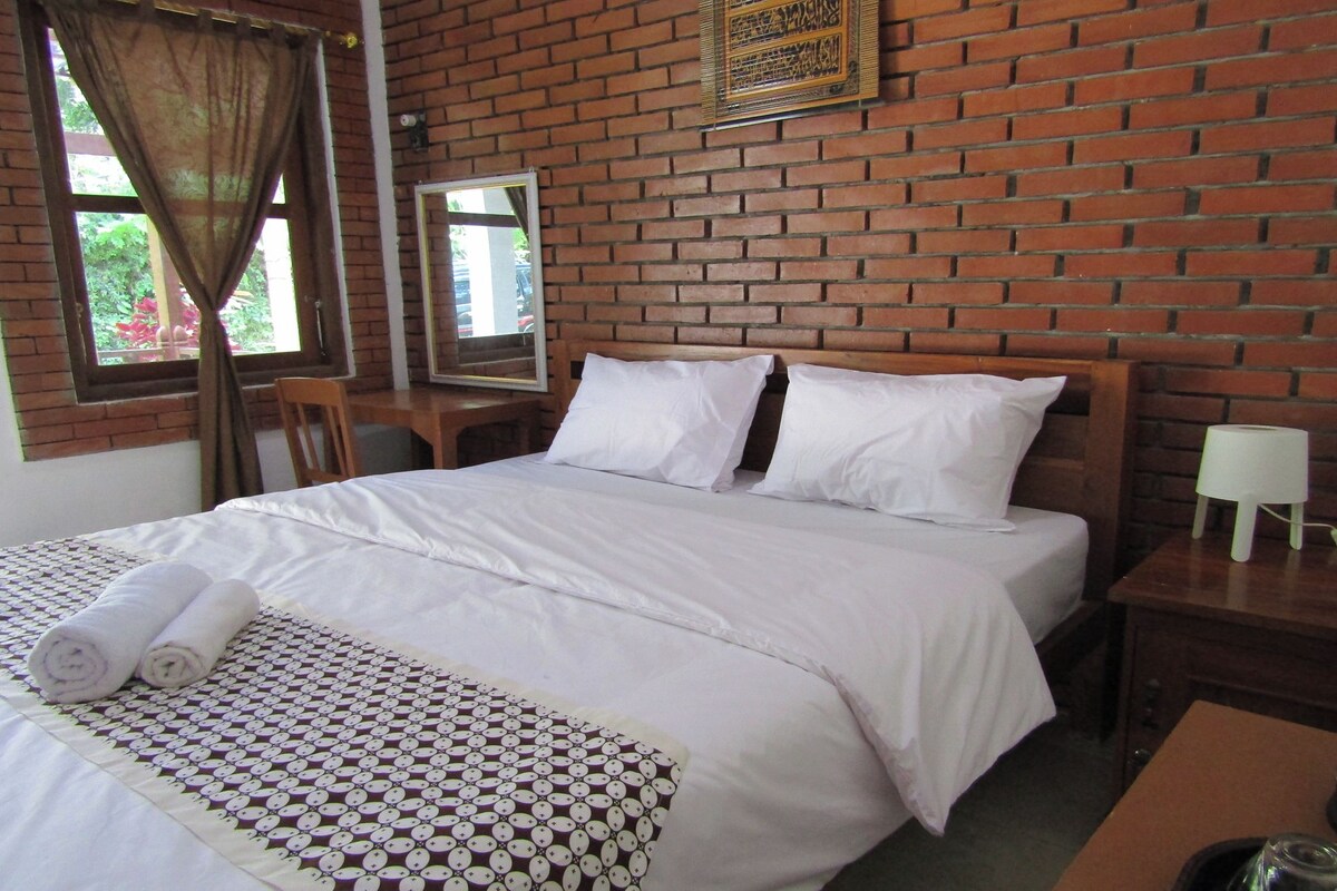 A hotel room with a brick wall, a large bed with white linens, a wooden nightstand, and a window with curtains.