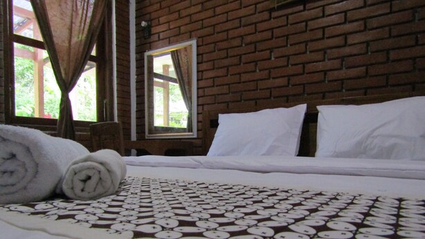 Cama bem arrumada com lençóis brancos e manta estampada, toalha enrolada e janela com cortinas em um quarto com paredes de tijolo.