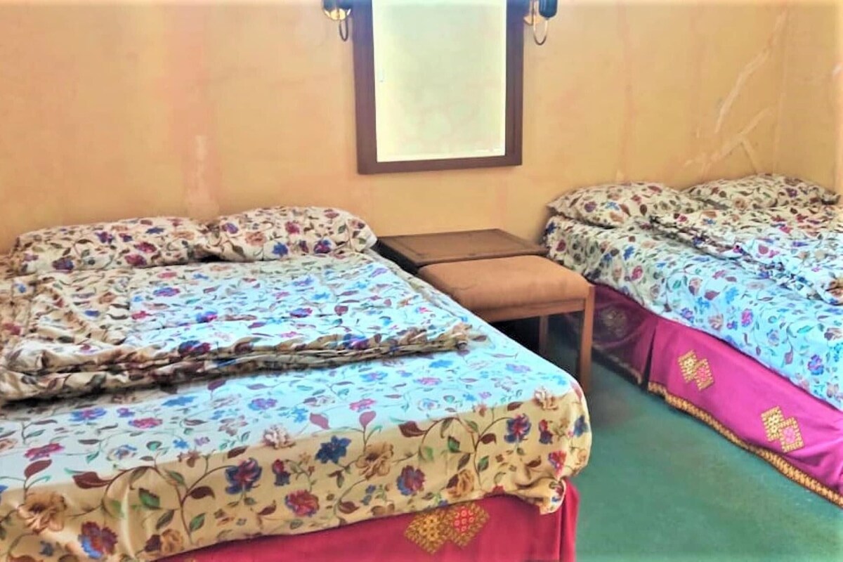 A room with two beds, each with floral bedding, a small wooden table, and a window with blinds.