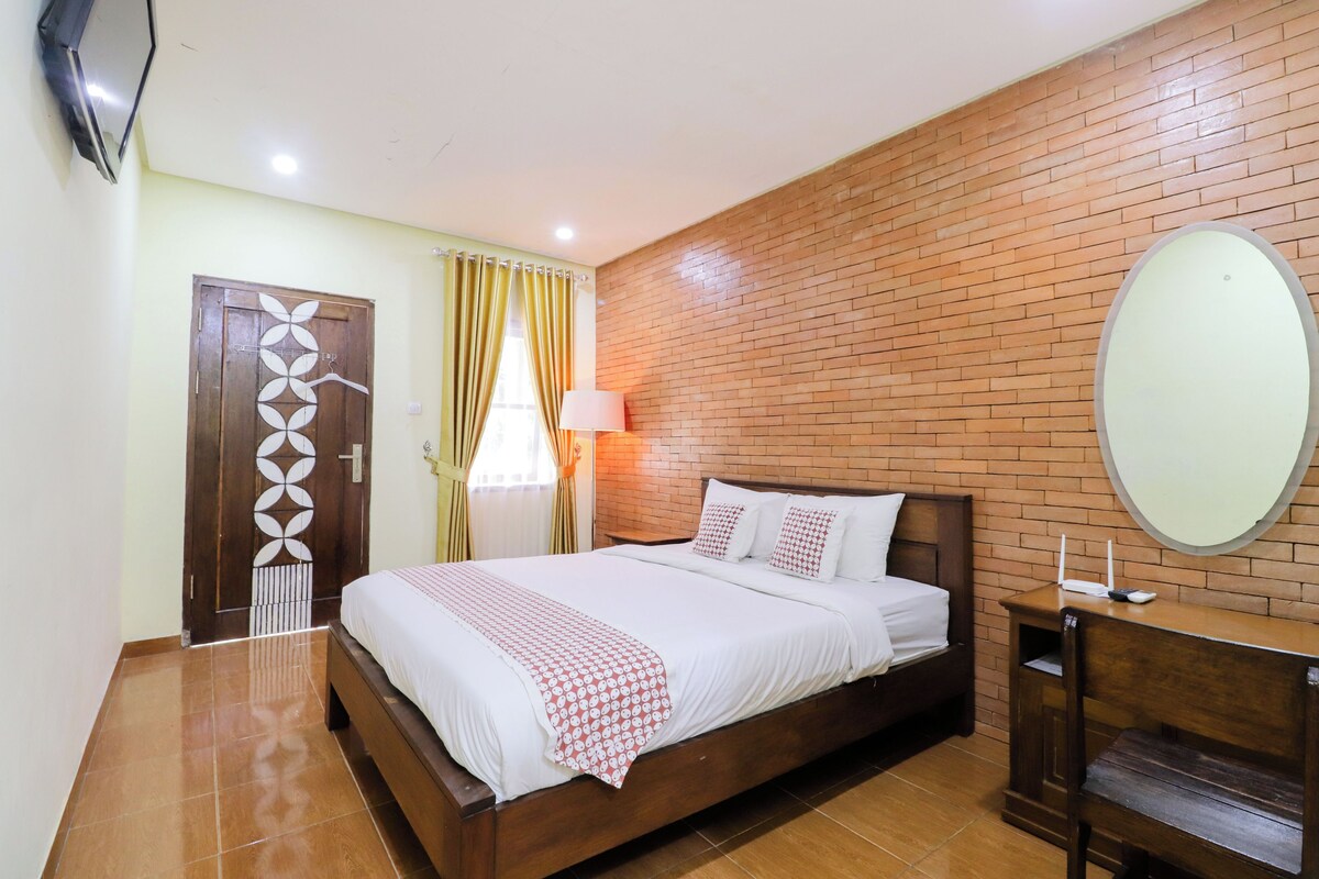 Una habitación de hotel con una cama de madera, una mesita de noche, un espejo, una pared de ladrillo, una puerta con panel decorativo y una ventana con cortinas.
