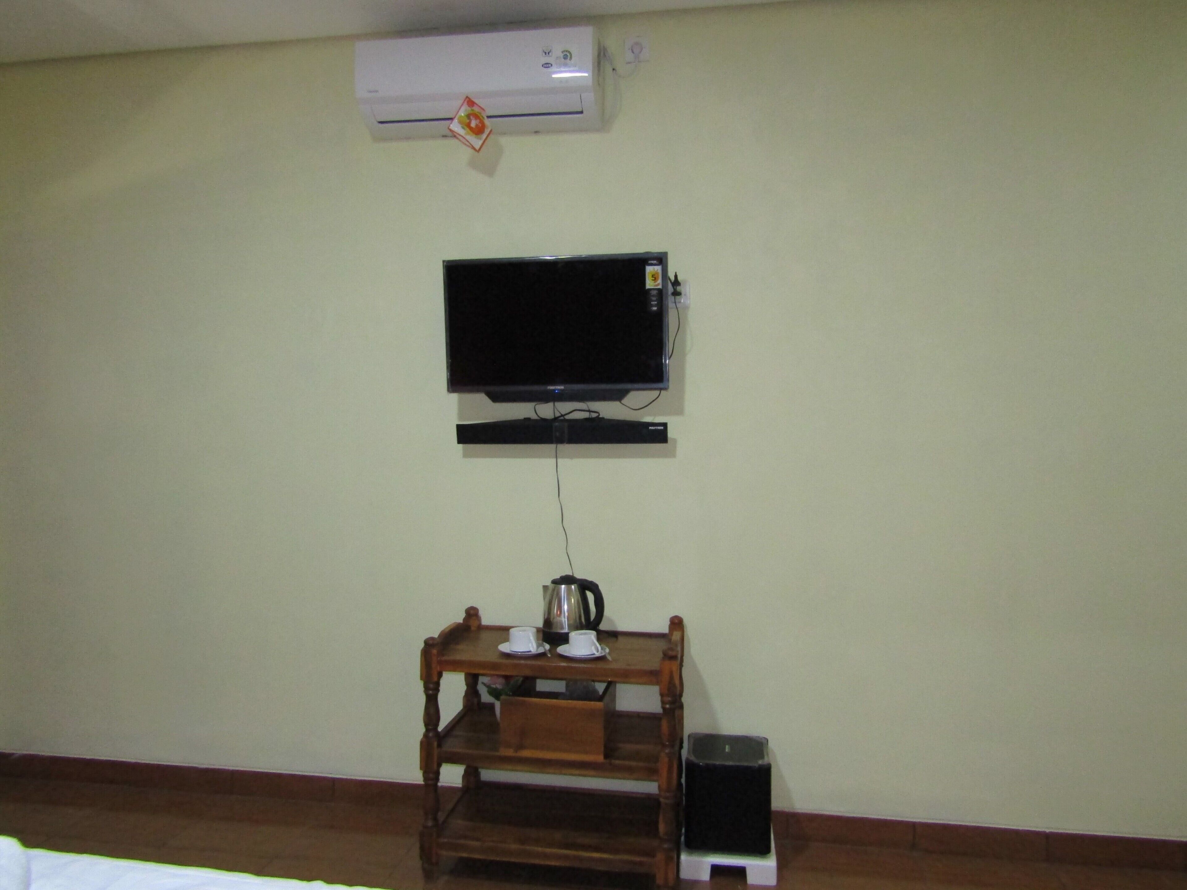 A room with a mounted flat-screen TV, a wooden table with a kettle, and a trash bin.