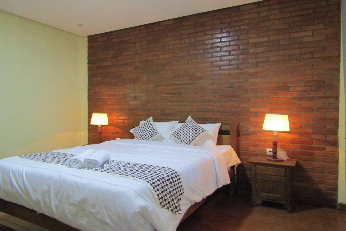 Een hotelkamer met een bakstenen muur, een bed met wit beddengoed, twee bedlampen en een keurig opgemaakt bed met handdoeken.