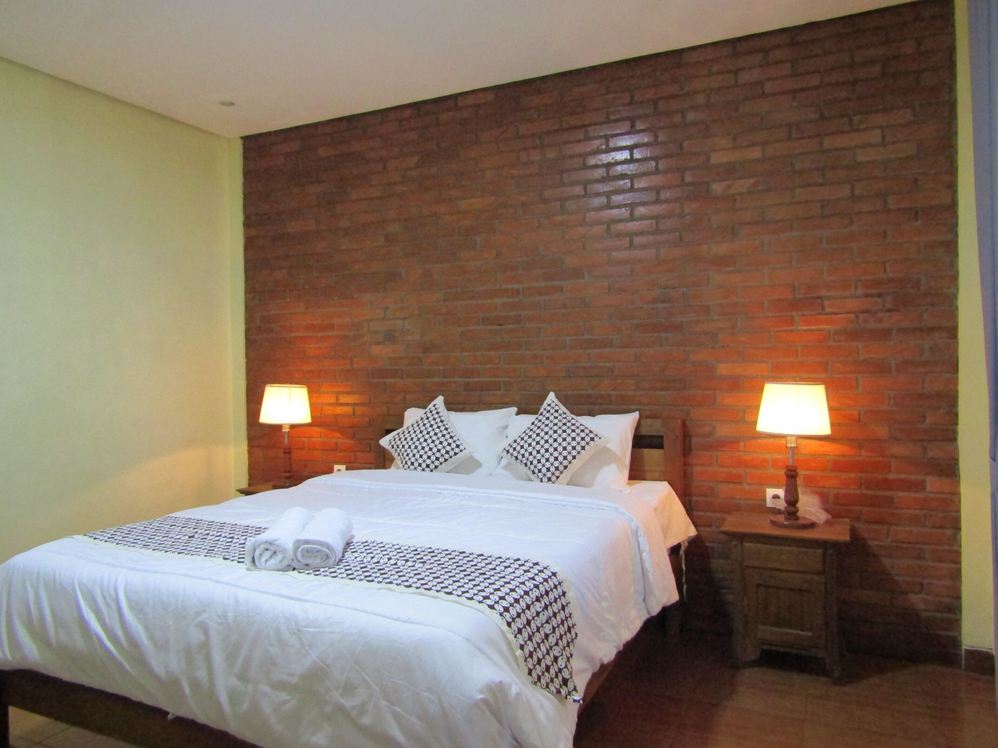 Une chambre d’hôtel avec un mur en briques, un lit bien fait avec du linge de lit blanc, deux tables de chevet avec des lampes et une petite table en bois.
