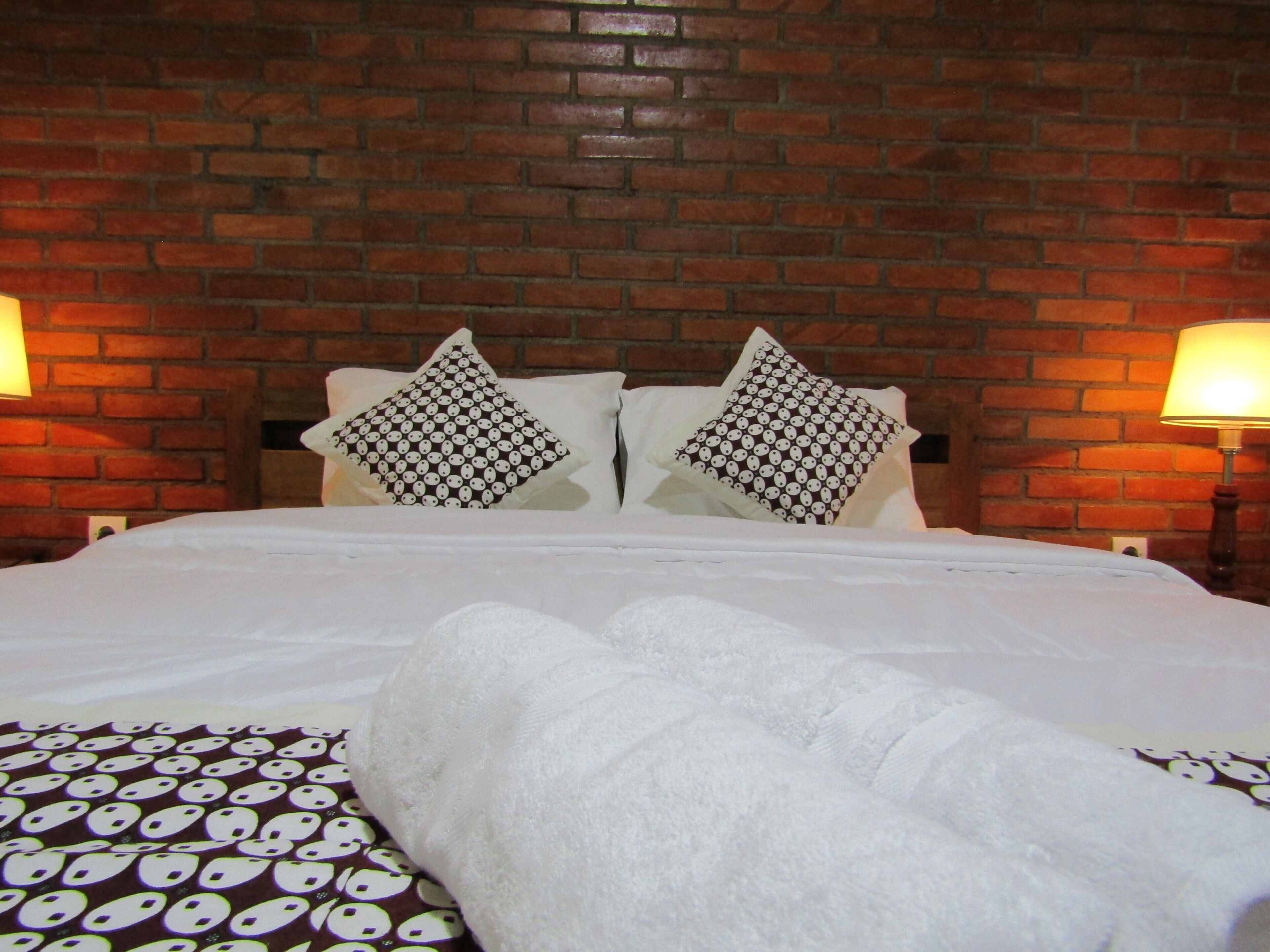 A hotel room with a brick wall, two bedside lamps, a bed with white bedding, and patterned pillows.
