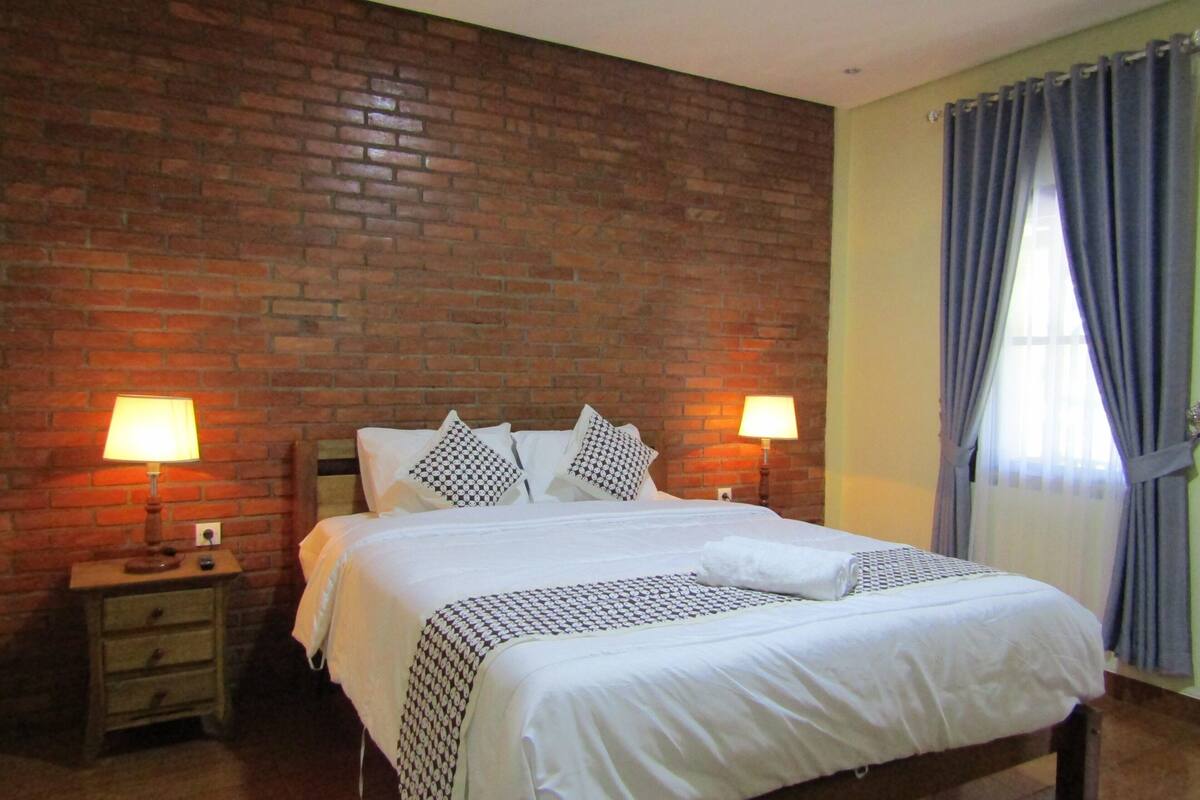 Un dormitorio con pared de ladrillo, cabecero de madera, dos mesitas de noche con lámparas y una ventana con cortinas.
