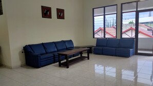 A spacious living room with two blue sofas, a wooden coffee table, and a large window offering a view of buildings outside. - Hotel Wina Wisata (Bandungan)