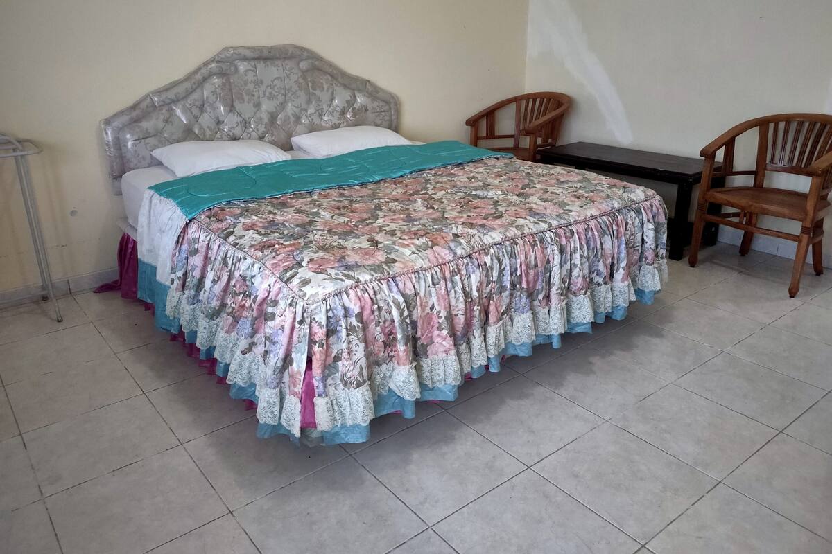 A bedroom with a bed, two wooden chairs, a small table, and a tiled floor.