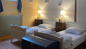 A room with two single beds, a wooden bench, a radiator, and two bedside tables with lamps and flowers.