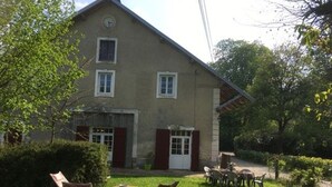 A two-story house with a front door, windows, and a clock on the wall. There is a small garden with grass, a bench, and a table with chairs.