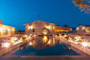 Zona de piscina con arquitectura de piedra, un edificio de piedra con veleta y una piscina bien iluminada con plantas alrededor.