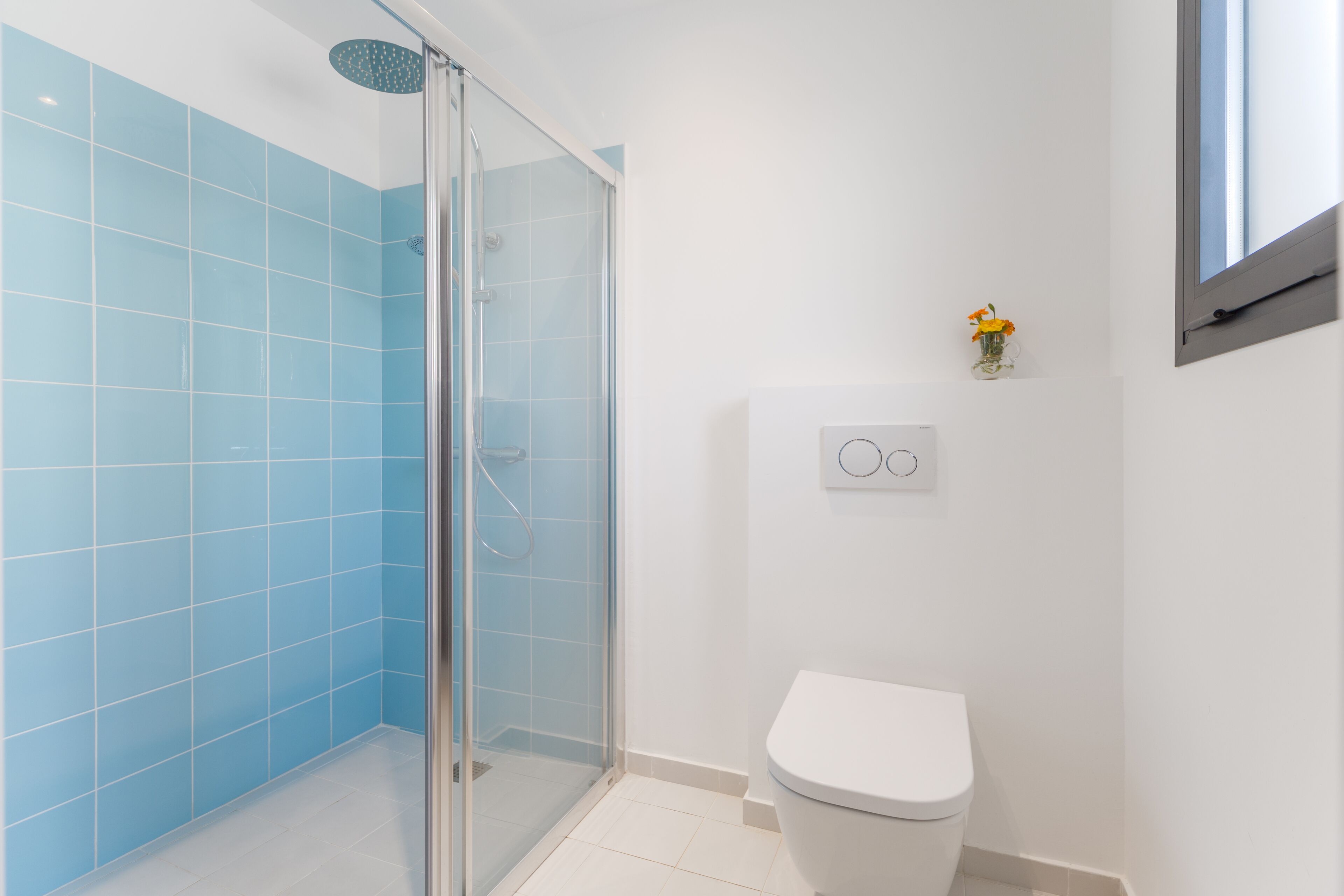 A bathroom with a glass shower enclosure, a white toilet, and a small vase with yellow flowers on the wall.