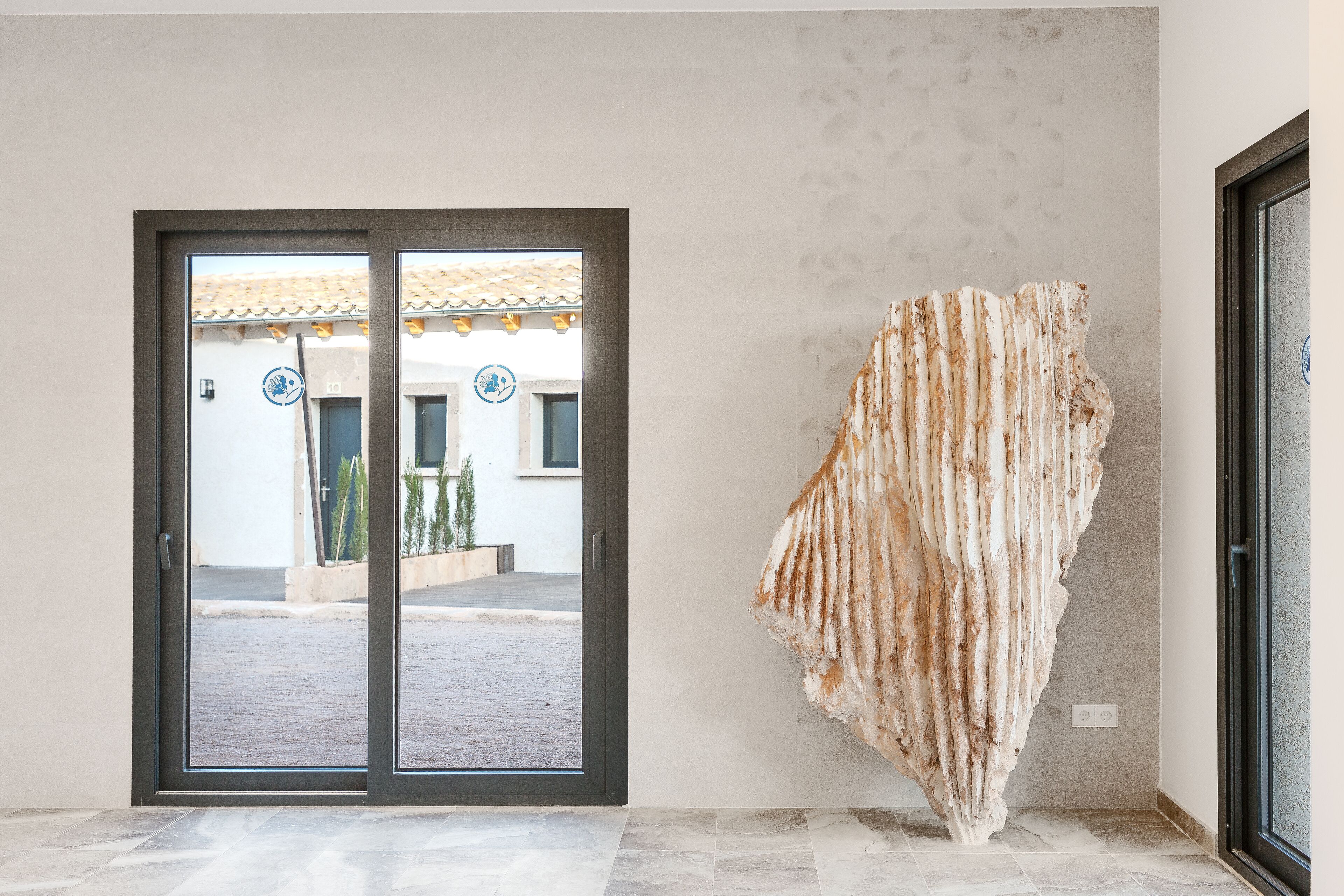 Una habitación con una gran escultura abstracta en la pared, un cerramiento de vidrio corredero que da a un espacio exterior y un suelo de baldosas.
