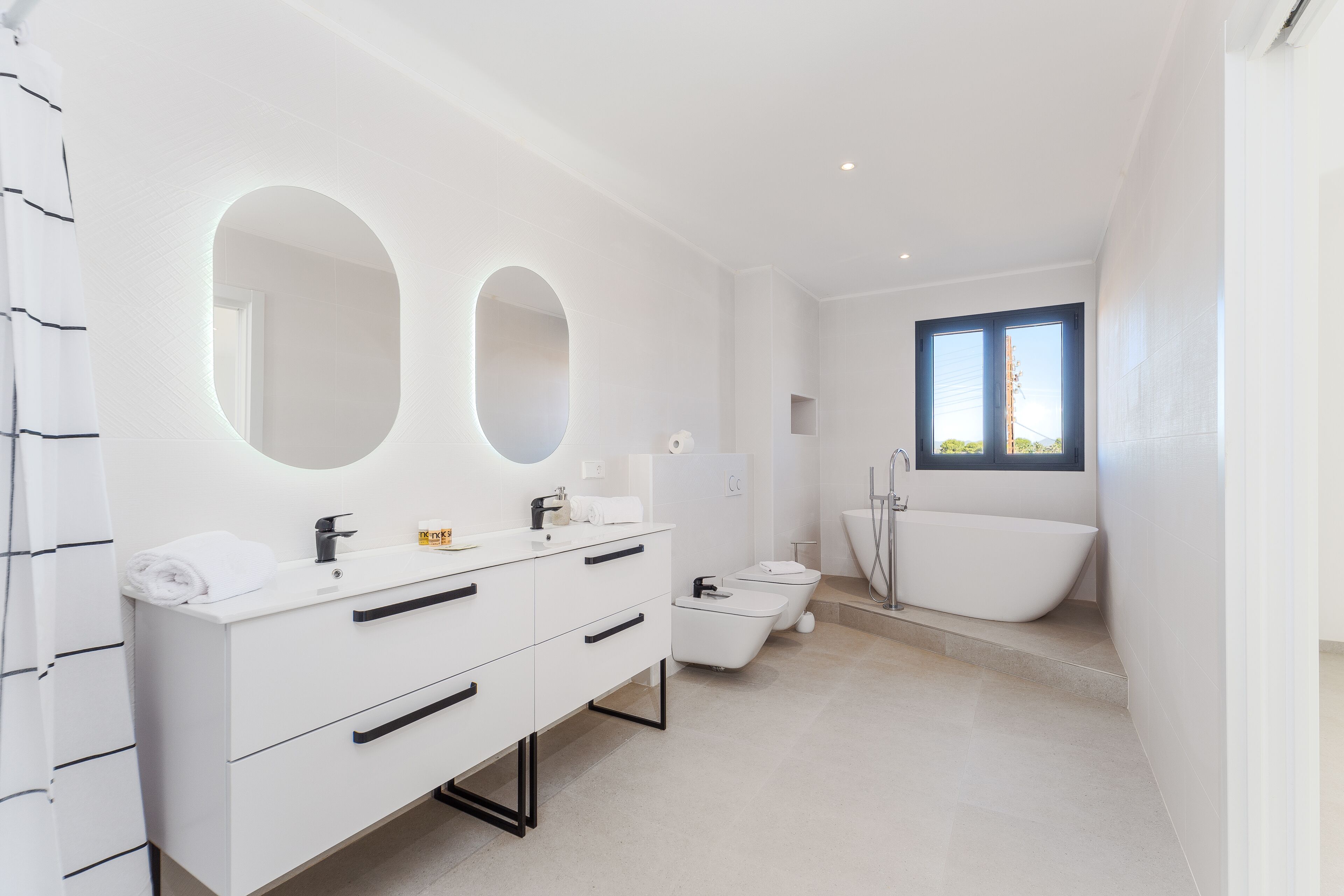 Un baño moderno con un tocador blanco, dos espejos ovalados, una bañera independiente y un inodoro.