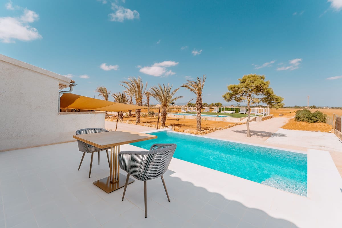 Un espace piscine extérieur moderne, doté d’une table et de chaises, entouré de palmiers et surplombé d’un ciel dégagé.