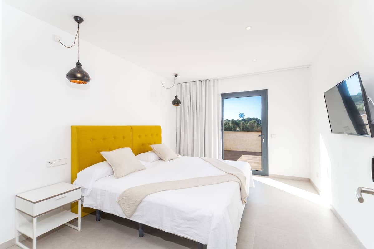 Une chambre à coucher moderne avec un grand lit, une tête de lit jaune, une table de chevet et un téléviseur fixé au mur.