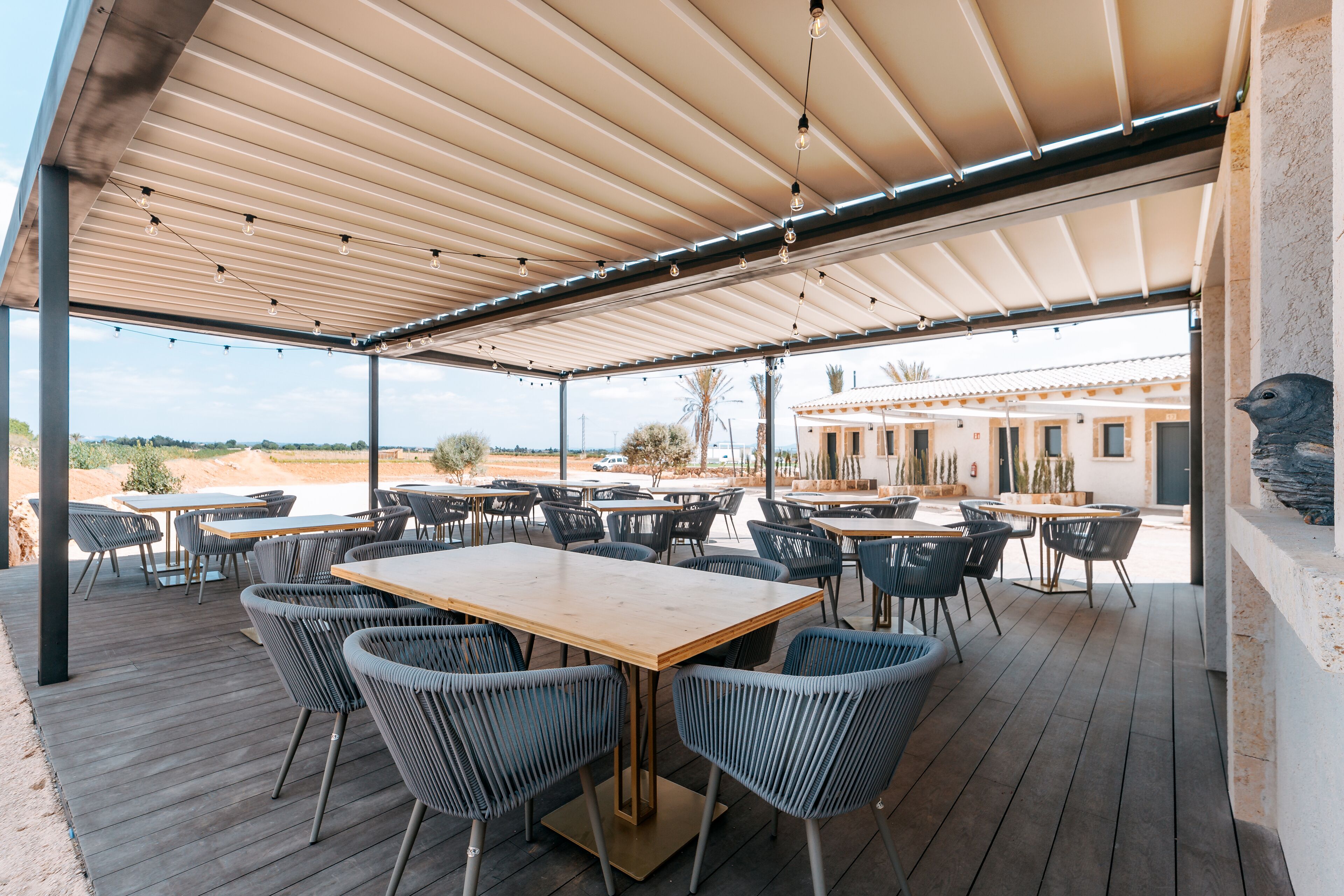 Un patio al aire libre con mesas y sillas de madera, una zona cubierta con techo a rayas y vistas a un edificio al fondo.