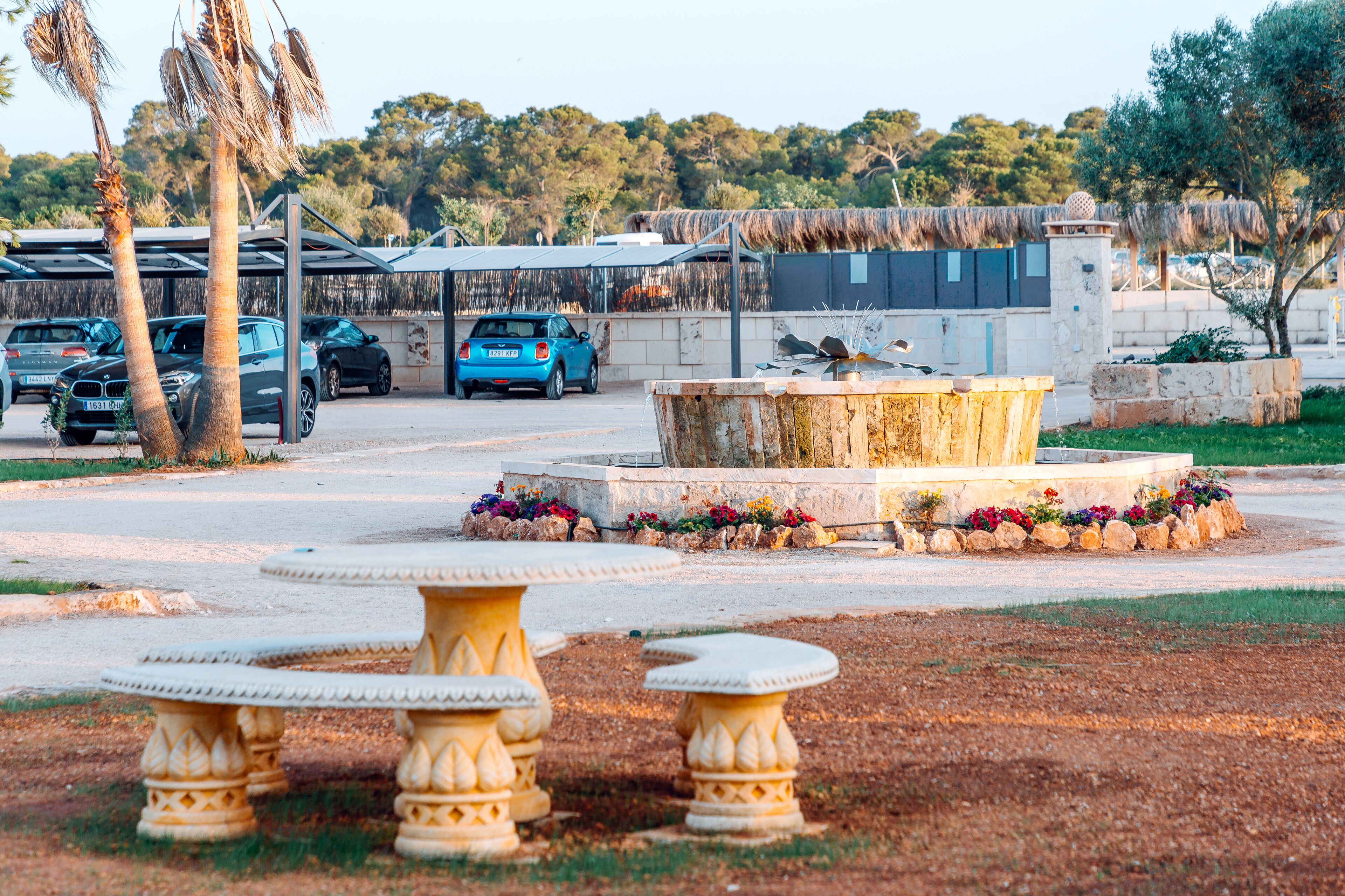 A courtyard with a central fountain, surrounded by stone tables and benches, with parked cars and a building in the background.