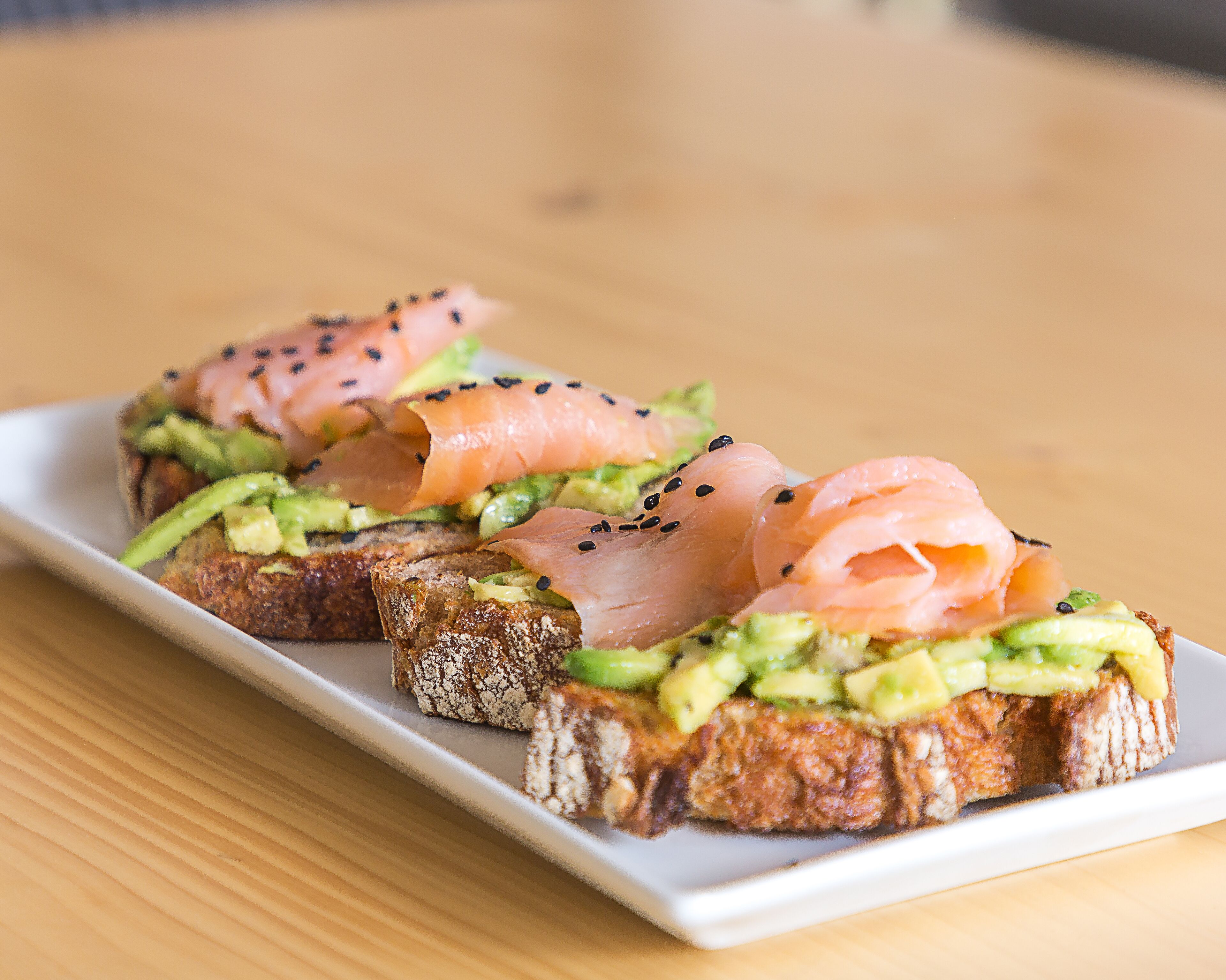 Un plato de comida con pan cubierto de aguacate y salmón ahumado, decorado con semillas de sésamo negro.