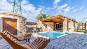 Zona de piscina con patio de piedra, sillones de madera y un pequeño rincón de estar al aire libre con mesa y sillas.