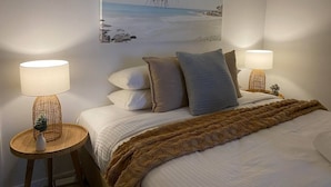 Una habitación de hotel con una cama, dos lámparas de noche, un cuadro de playa y una mesita redonda con un florero.