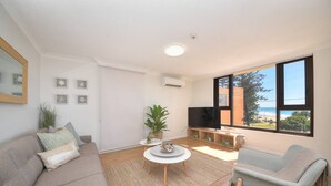 Una sala de estar moderna con sofá, sillón y mesa de centro. Hay un televisor sobre un mueble bajo y un ventanal con vistas a la playa.
