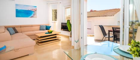 A modern living room with a sofa, a coffee table with fruits, a TV, and a balcony with a table set for dining.