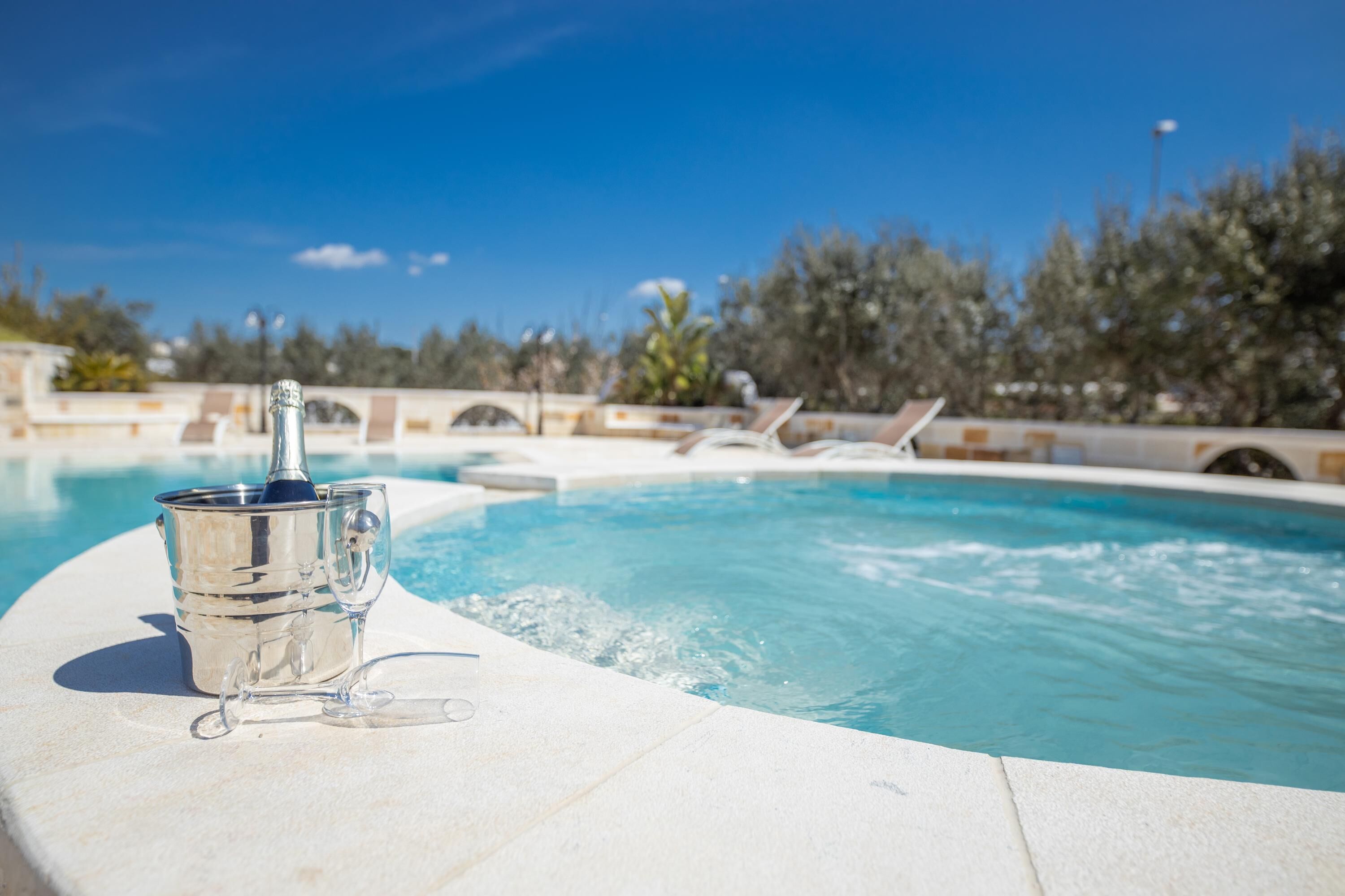 Un área junto a la piscina con un cubo metálico, un vaso, sillas de descanso y un cielo azul despejado.