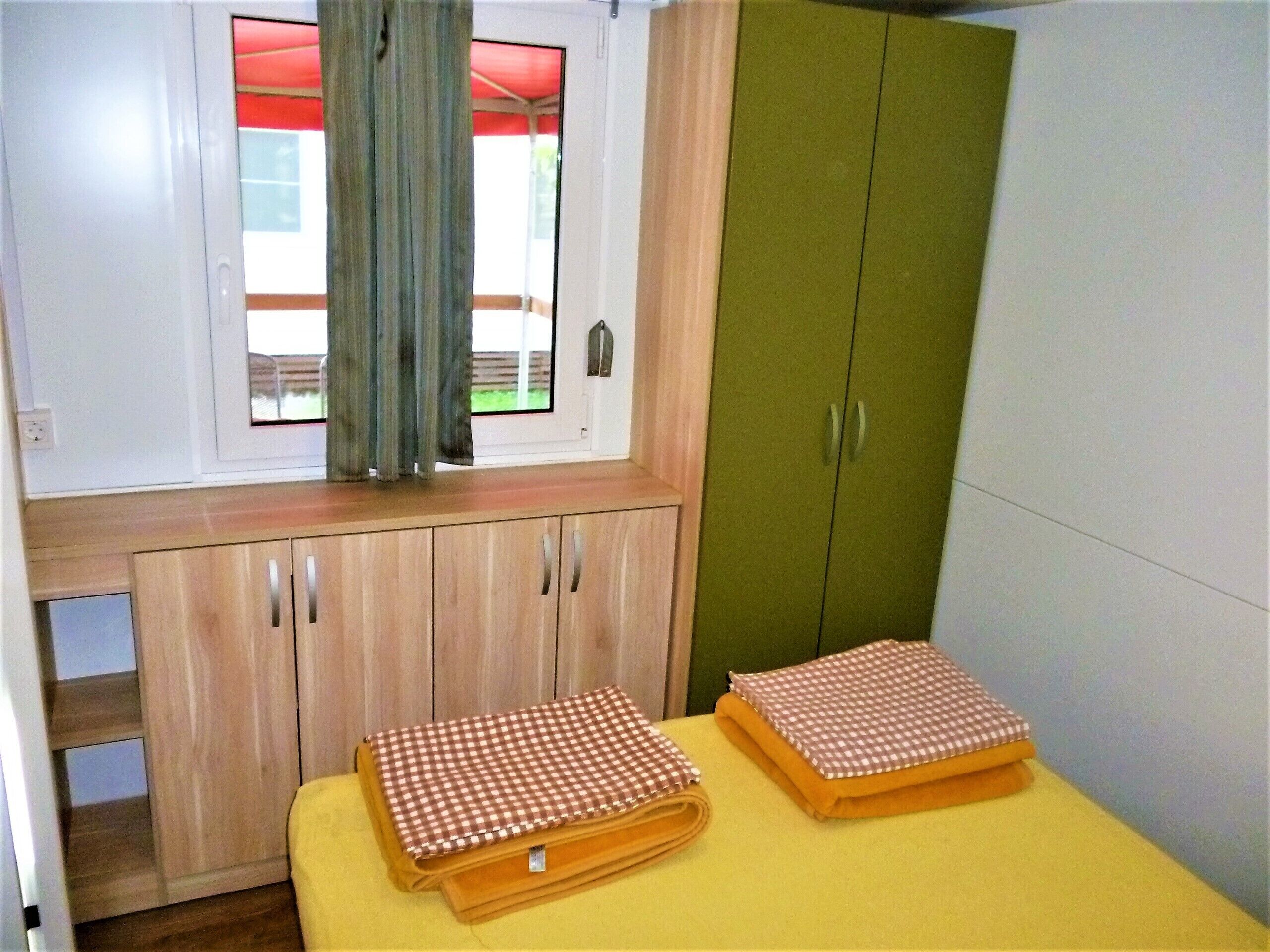 Una habitación pequeña con ventana, un armario verde y una mesa amarilla con manteles a cuadros.