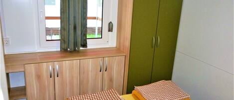 Una habitación pequeña con ventana, un armario verde y una mesa amarilla con manteles a cuadros.