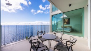 Un balcon avec une table et des chaises, une porte vitrée donnant sur une chambre, et une vue sur l’océan.