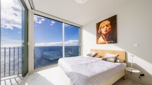 Une chambre avec une grande fenêtre, un lit avec des oreillers, une table de chevet et une affiche accrochée au mur.