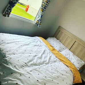 A bed with white bedding featuring a bee pattern, a wooden headboard, and a window with a view of a green lawn and a building. - Charming 3-bed Holiday Home at Primrose Valley (Filey)
