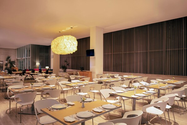 A spacious dining area with multiple tables and chairs, a large hanging light fixture, and a television screen on the wall. - Fiesta Inn Morelia Altozano (Morelia)