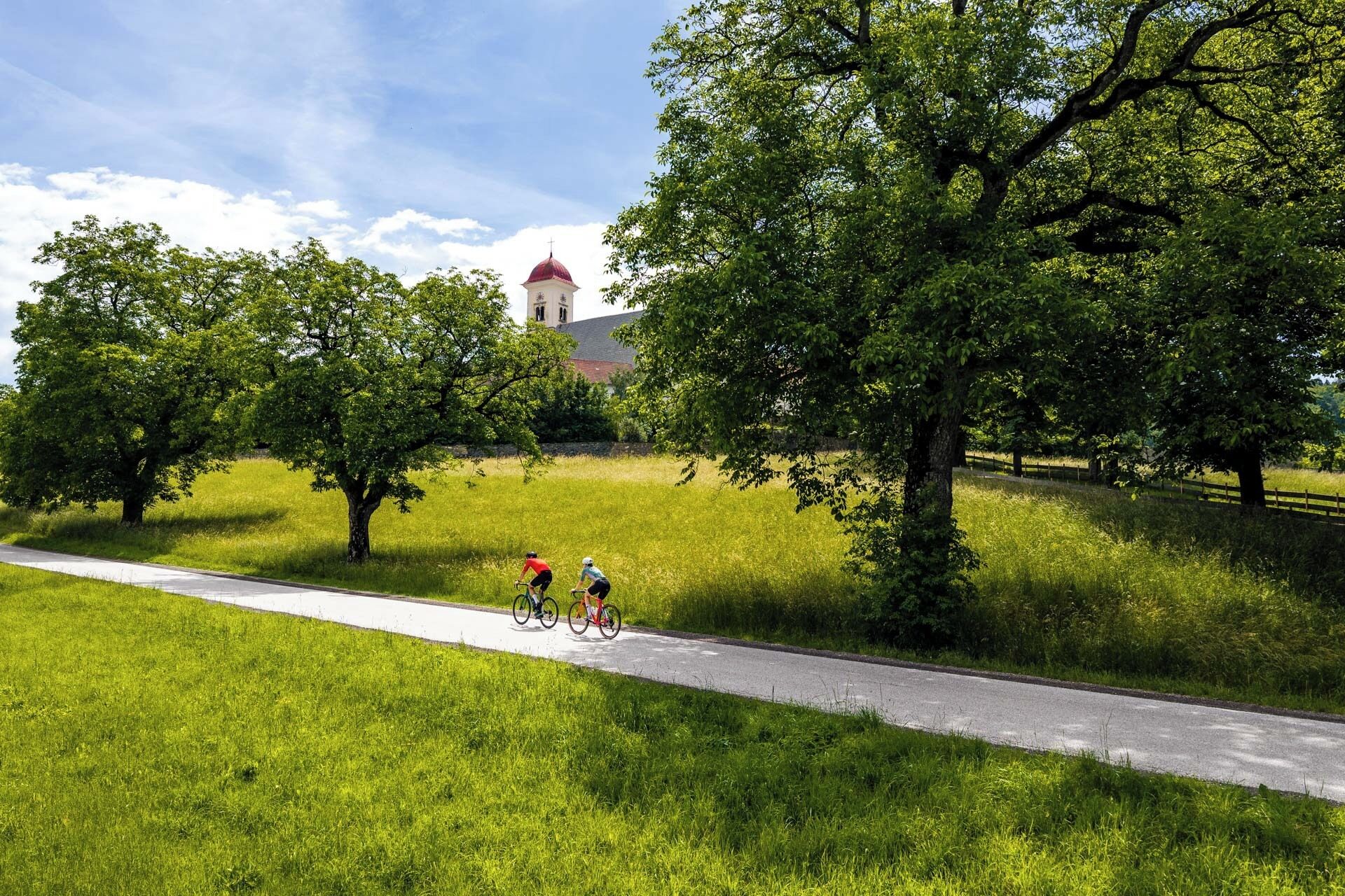 Un chemin pavé où l’on voit des cyclistes, entouré d’une végétation luxuriante, avec un bâtiment à toit rouge en arrière-plan.