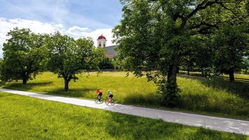 A paved path with cyclists, surrounded by lush greenery and a building with a red roof in the background.