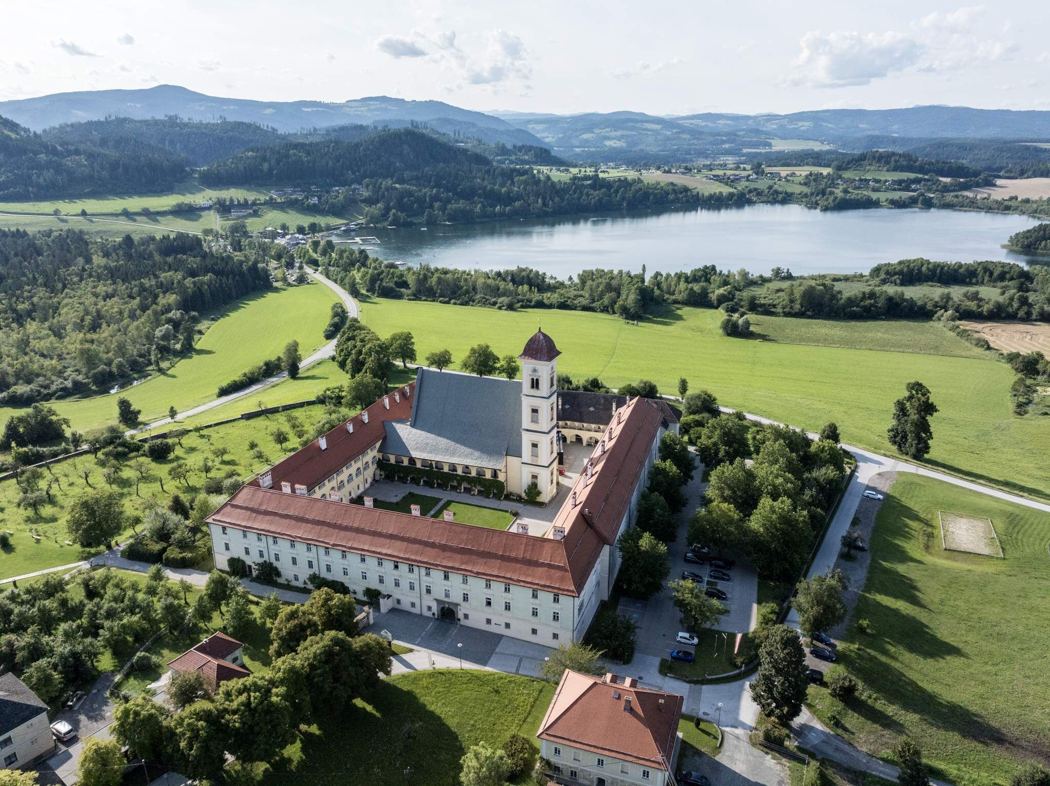 A large, historic building with a central courtyard, surrounded by lush green fields and a lake.