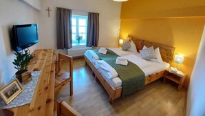 Une chambre d’hôtel avec un lit, une table de chevet, une lampe, une télévision, une fenêtre avec des rideaux et une croix accrochée au mur.